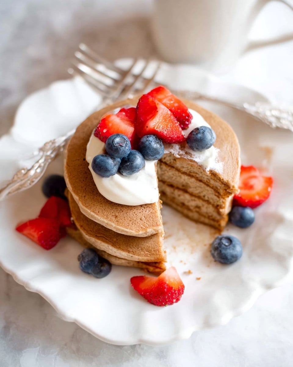 A stack of three light brown pancakes sits on a white plate with scalloped edges, placed on a white marbled surface. The top pancake has a bite taken out of it, showing its soft, fluffy inside. On top, there is a dollop of white cream, with fresh blueberries and small pieces of red strawberries scattered on and around the stack. A silver fork rests on the plate behind the pancakes, and in the background, there is a white cup partially visible. photo taken with an iphone --ar 4:5 --v 7