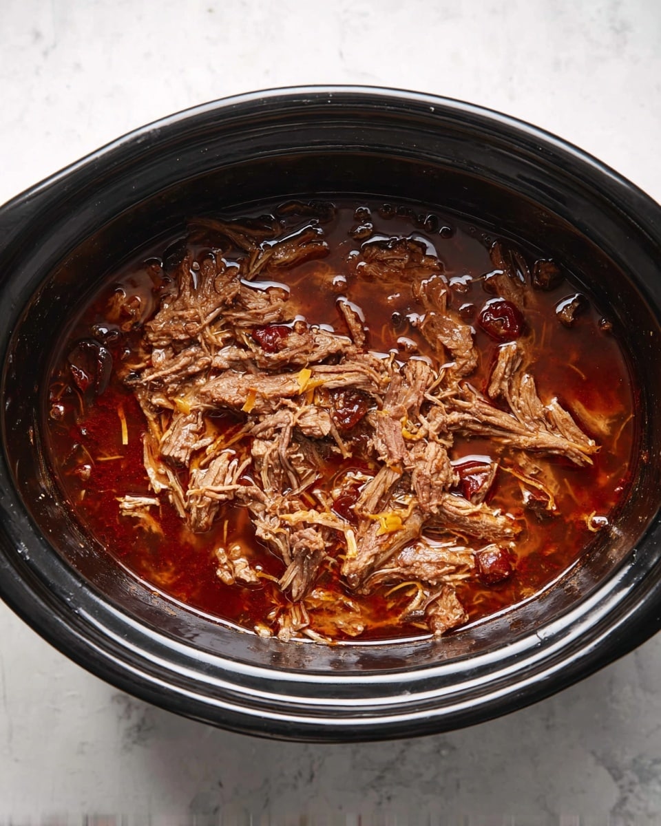 A black slow cooker filled with shredded cooked meat in a reddish-brown broth with visible pieces of chili and thin strands of onion or garlic. The meat has a tender texture, mixed evenly with the sauce, and small bits float throughout the liquid. The slow cooker is placed on a white marbled surface. photo taken with an iphone --ar 4:5 --v 7