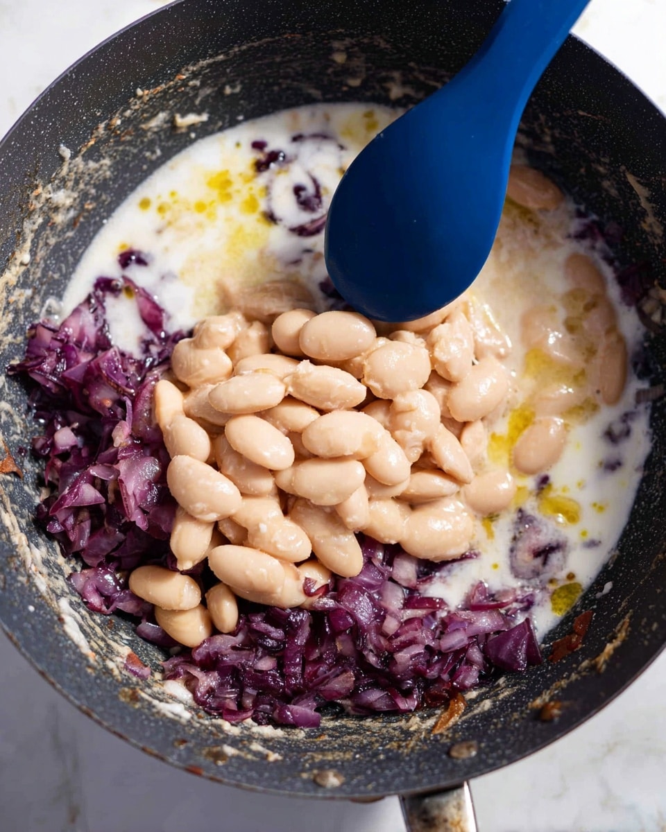 A close-up view of a black pan with a layer of cooked chopped red onions at the bottom, showing a dark purple color and soft texture. On top of the onions, there is a creamy white liquid mixed with small pools of yellow oil. Over this, there is a large layer of light beige beans with smooth, shiny surfaces, being stirred by a blue spoon in the middle of the pan. The pan edges and base show speckles and food residue. The background is a white marbled texture. Photo taken with an iphone --ar 4:5 --v 7