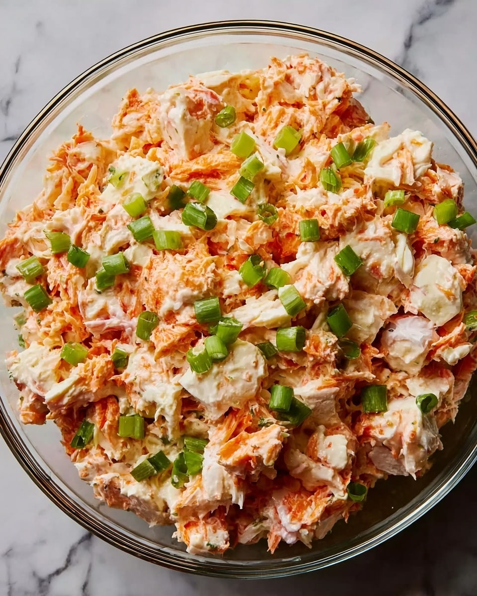 A clear glass bowl filled with a mix of orange and white crab pieces, coated in a creamy light orange sauce, with small bright green chopped scallions scattered on top. The crab pieces vary in size and texture, some shredded and others in chunks. The bowl sits on a white marbled surface. photo taken with an iphone --ar 4:5 --v 7