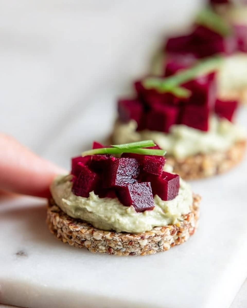 The image shows three round crackers placed in a vertical line on a white marbled surface. Each cracker has a base layer of light brown seeds and grains with a rough texture. On top of each cracker, there is a thick layer of pale green creamy spread evenly covering the surface. The final layer consists of small, bright red cubes of beetroot piled in the center, topped with thin, short green chive pieces. A woman's hand is slightly visible holding the edge of the middle cracker. The focus is sharp on the middle cracker, with the others softly blurred in the background. Photo taken with an iphone --ar 4:5 --v 7
