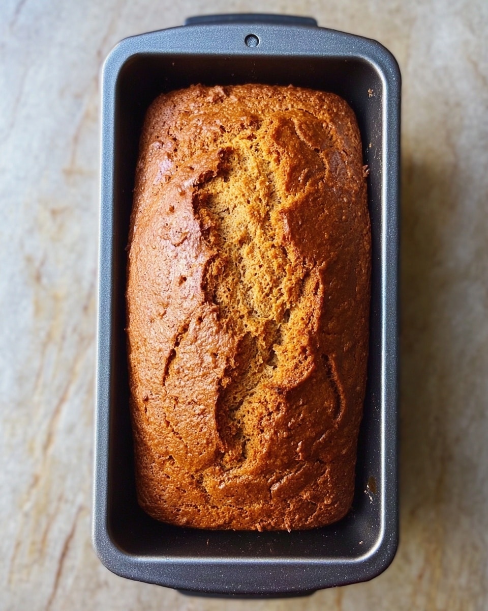 A close-up top view of a freshly baked brown loaf in a gray metal baking pan. The loaf has a rough, cracked surface showing a golden-brown color with a soft texture. The baking pan is placed on a white marbled surface, and the loaf fills the pan fully with slightly rounded edges. photo taken with an iphone --ar 4:5 --v 7