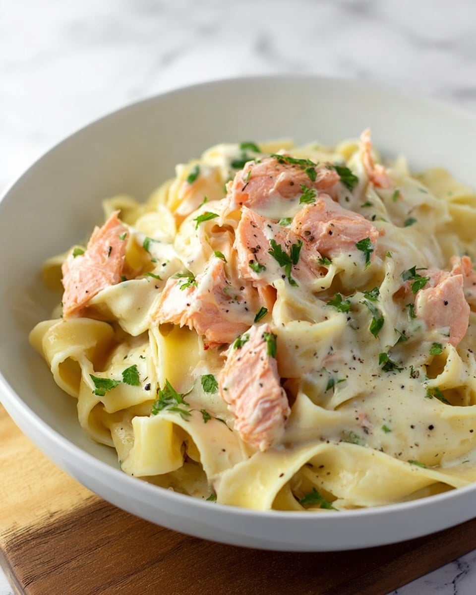 The dish shows creamy fettuccine pasta layered at the bottom with smooth, thick white sauce coating it evenly, topped with several pieces of pink cooked salmon scattered across the pasta, each piece flecked slightly with black pepper. Small bright green parsley leaves are sprinkled on top, adding color contrast. The food is served in a white bowl placed on a white marbled surface, with soft natural light highlighting the textures and colors. photo taken with an iphone --ar 4:5 --v 7