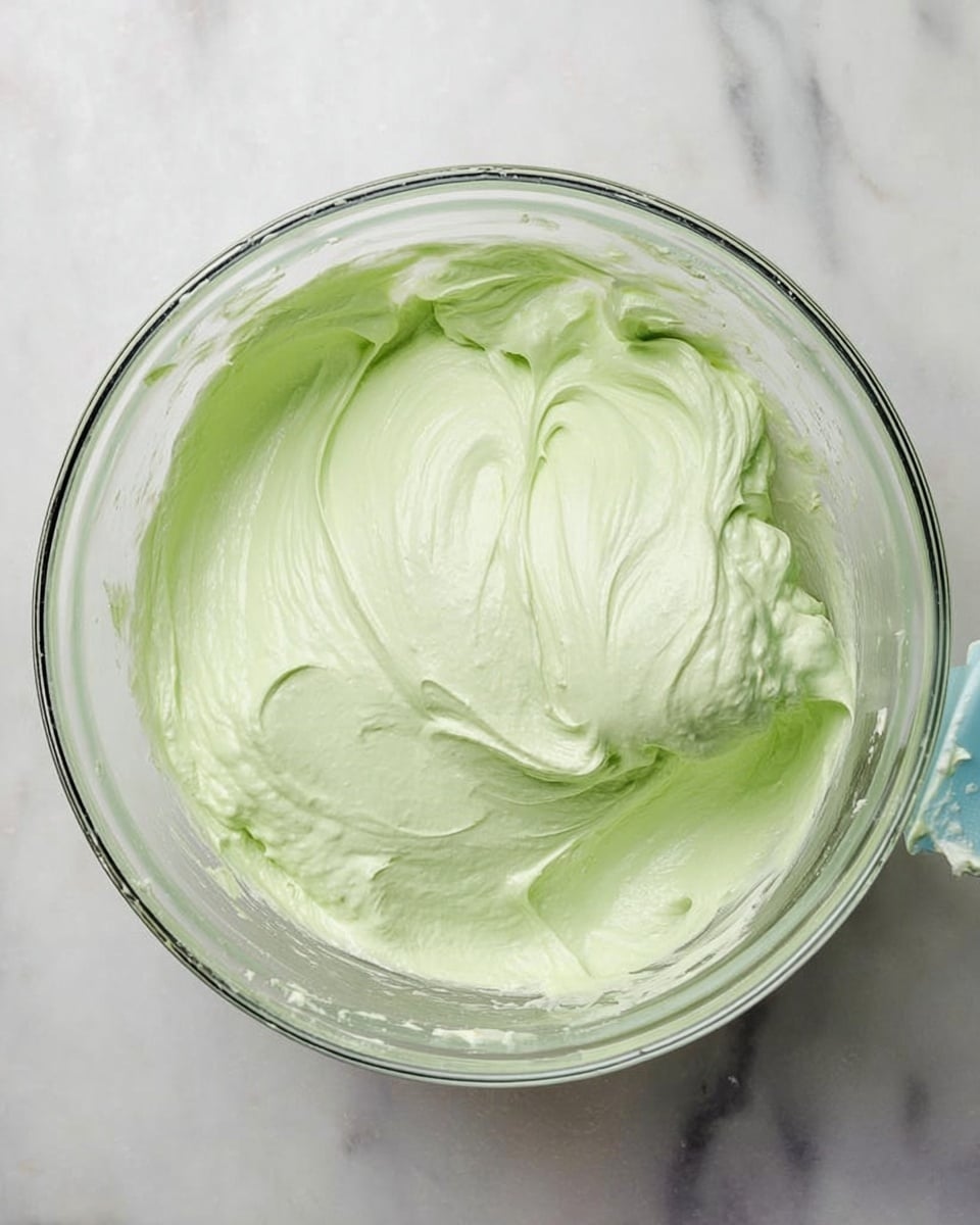 A clear glass bowl is filled with a smooth, light green creamy mixture that looks soft and fluffy. The cream has a slightly glossy texture, with visible soft peaks and folds showing where it has been mixed or stirred. The bowl sits on a white marbled surface, and a hint of a blue mixing tool can be seen at the bowl’s edge. photo taken with an iphone --ar 4:5 --v 7