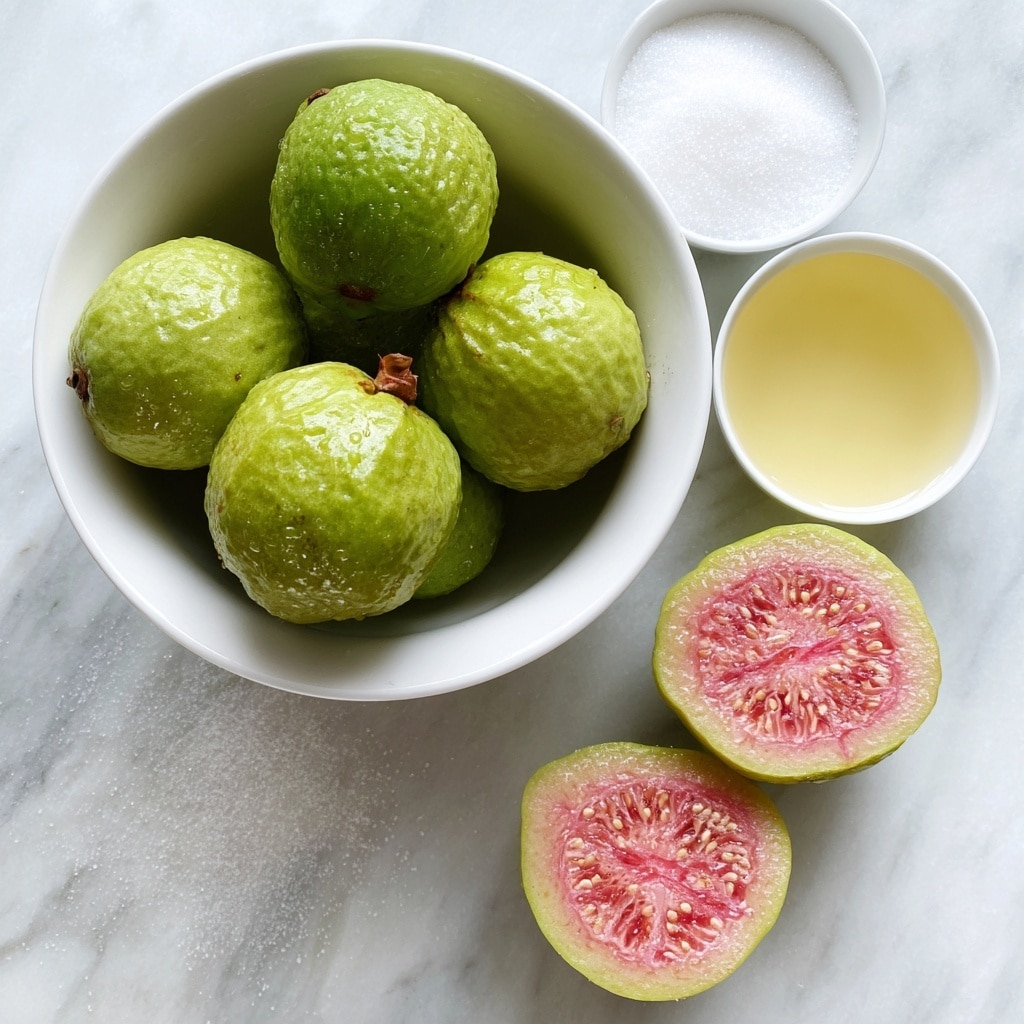 A white bowl filled with whole green guavas and two guavas cut in half showing their pink flesh and seeds is placed at the bottom right of the image. Above the bowl on the left side, there is a small white bowl filled with white sugar, and on the right side, another small white bowl contains light yellow lime juice. The background is a white marbled texture with the words