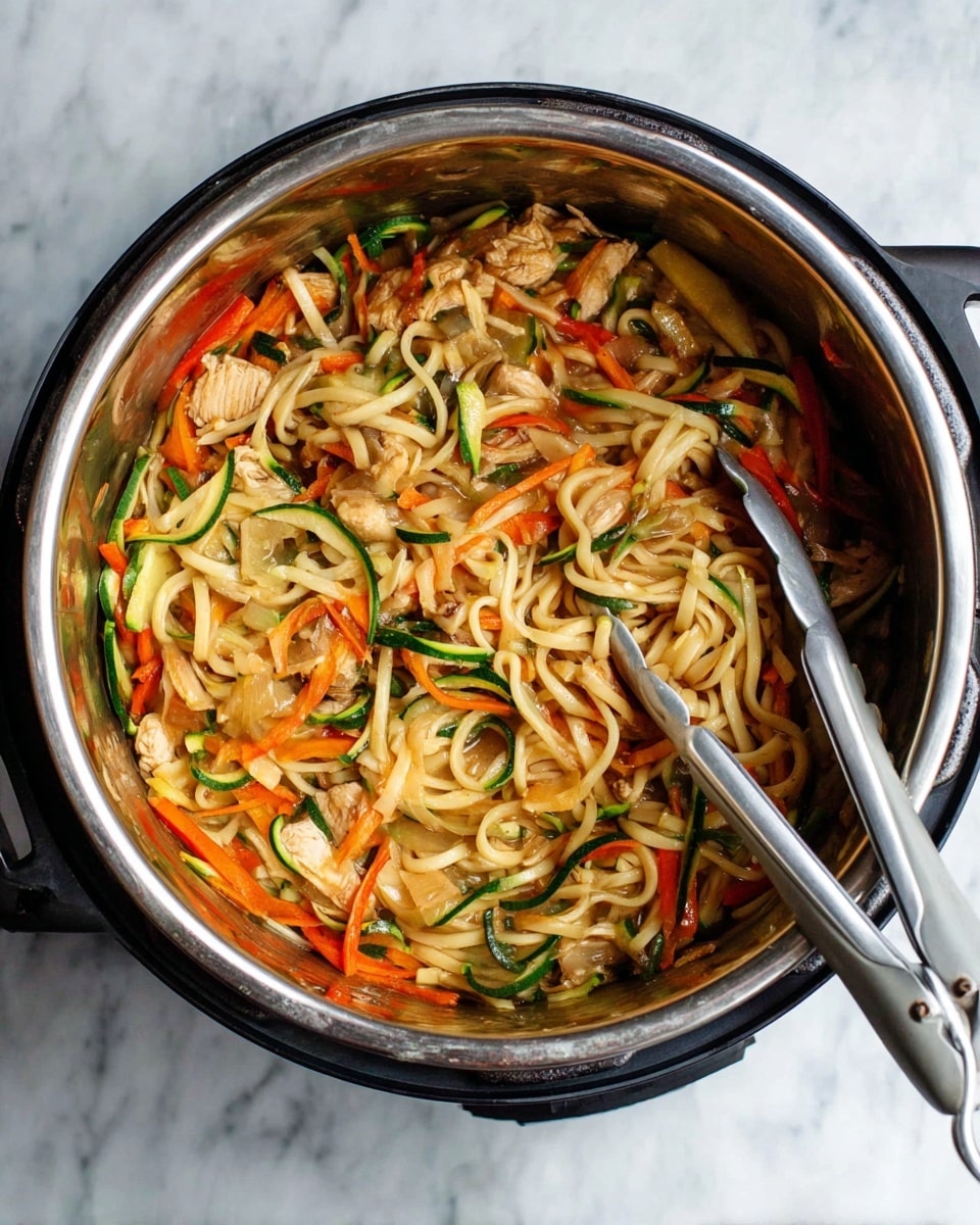 A stainless steel pot filled with cooked noodles mixed with sliced orange carrots, light green zucchini strips, and light brown chicken pieces. The noodles are light beige and thick, blending with the colorful vegetables inside the pot. A gray and silver tong rests inside, holding some noodles. The pot is placed on a white marbled surface. photo taken with an iphone --ar 4:5 --v 7