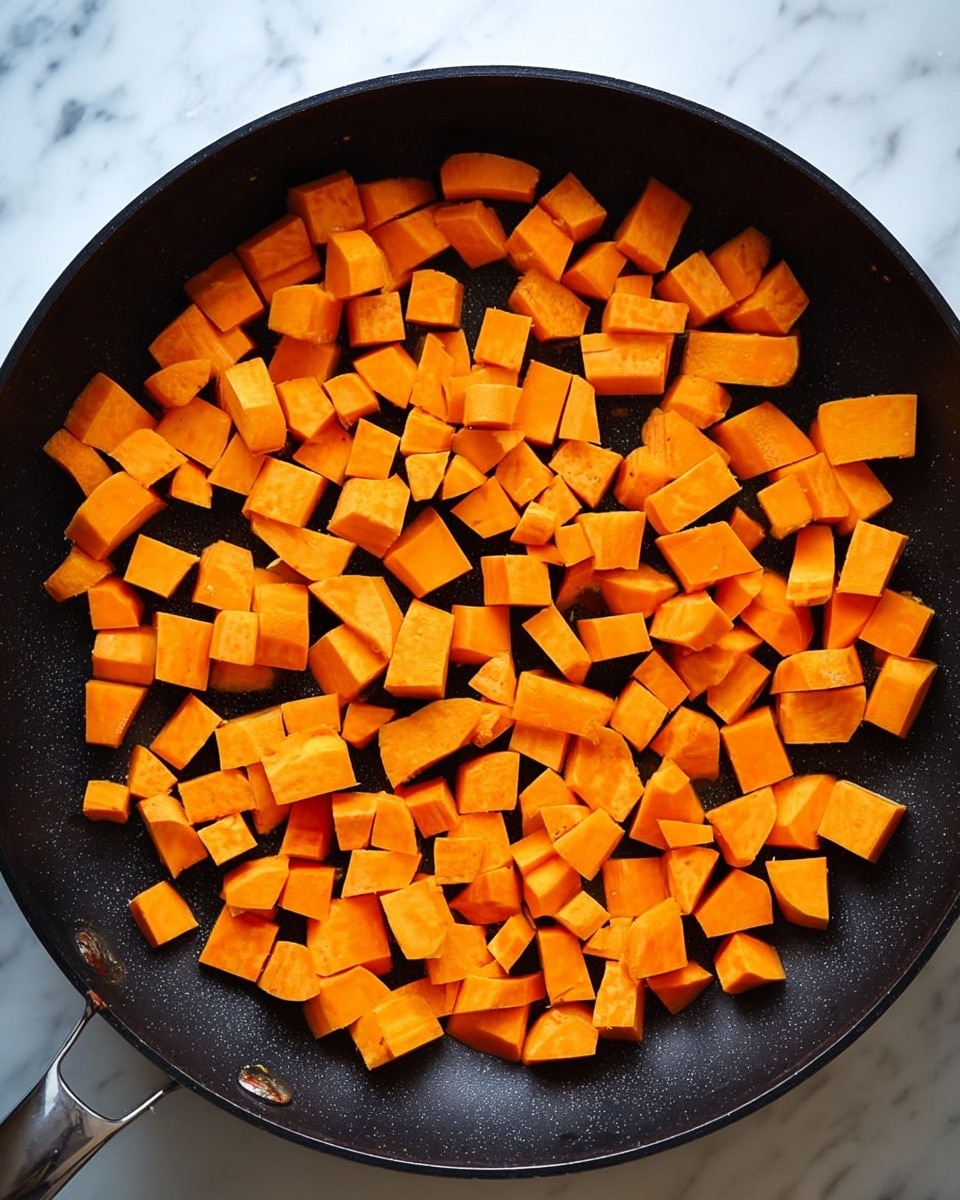 A black frying pan is shown from above filled with many small, cubed pieces of bright orange sweet potato spread out evenly across the pan’s surface. The sweet potato pieces vary slightly in size and shape but mostly have a smooth texture and appear fresh and raw. The pan is placed on a white marbled surface. photo taken with an iphone --ar 4:5 --v 7