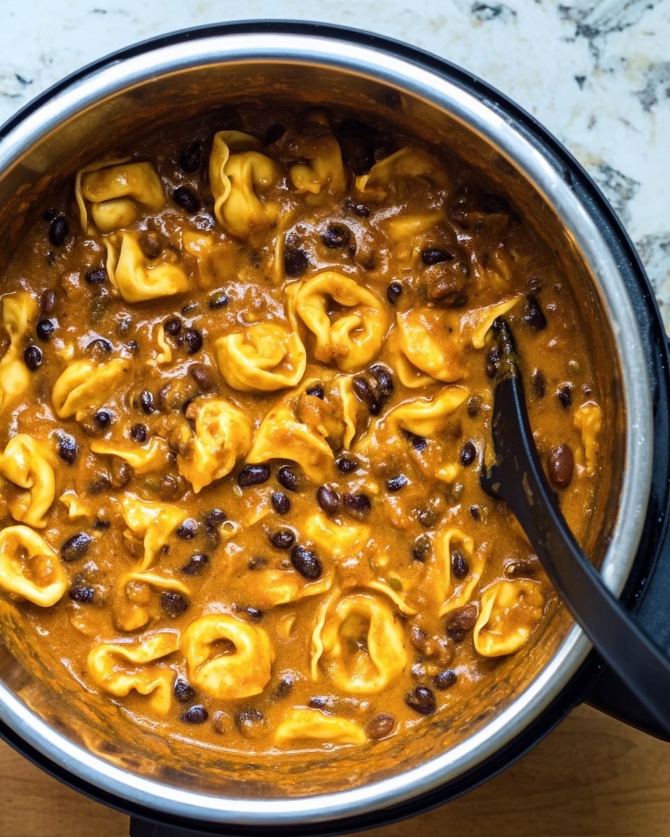 The image shows a close-up top view of a pot filled with a thick dish made of tortellini pasta and black beans in a rich orange sauce. The tortellini are light yellow with a soft, smooth texture, scattered evenly throughout the sauce. The black beans are small, shiny, and mixed well in the dish, adding contrast to the orange sauce and pasta. There is also a black spoon inside the pot, partially dipped in the food, visible on the right side. The pot is silver with a black rim, sitting on a wooden surface changed to white marbled texture. Photo taken with an iphone --ar 4:5 --v 7