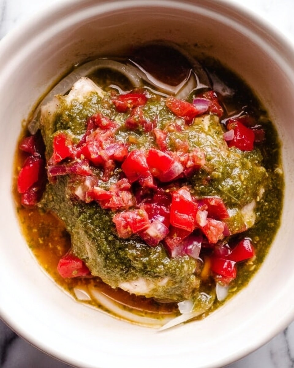 The image shows a white bowl with a layer of white onion slices at the bottom. On top, there are two chicken pieces mostly covered by thick green salsa with a chunky texture. Bright red chopped tomatoes are scattered over the green salsa, adding bright patches of color around the chicken. The dish is placed on a white marbled surface. photo taken with an iphone --ar 4:5 --v 7