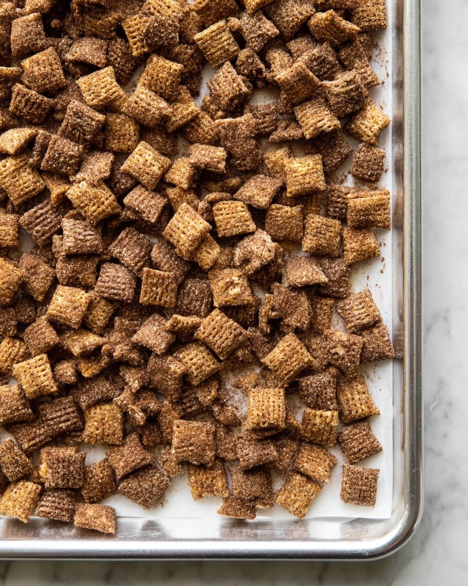 The image shows a single layer of square cereal pieces spread evenly on a baking sheet lined with white parchment paper. The cereal pieces are light brown with visible ridges and a rough texture. Each piece is coated in a darker brown powder, giving them a dusted, grainy look that covers the surface. The baking sheet has metal edges and the background surface features a white marbled texture. photo taken with an iphone --ar 4:5 --v 7