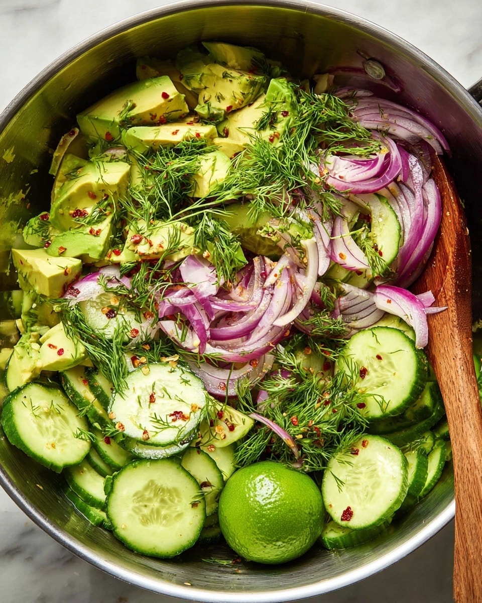 The image shows a metal bowl filled with a fresh salad made of several layers of ingredients: at the bottom are thinly sliced green cucumbers with light seeds, followed by chunks of bright green avocado, thin slices of light purple-red onion, and bright green fresh dill scattered on top. There are also some red chili flakes sprinkled over the salad, adding small red spots throughout. A wooden spoon is partly visible on the right side of the bowl, and a bright green lime resting in the bowl adds a fresh touch. The whole scene sits on a white marbled surface. Photo taken with an iphone --ar 4:5 --v 7