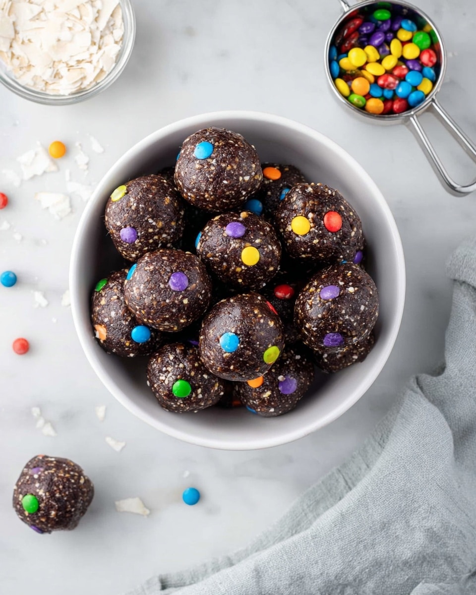 A white bowl is filled with about twelve dark brown round energy balls that have a rough texture and are dotted with small colorful candy pieces in red, blue, green, yellow, purple, and orange. The bowl sits on a white marbled surface, with a small metal measuring cup filled with more colorful candy pieces near the top right corner, and another measuring cup with white flakes near the bottom left. There are a few loose candy pieces scattered around the bowl, and a light gray cloth is placed on the bottom right side of the image. Photo taken with an iphone --ar 4:5 --v 7
