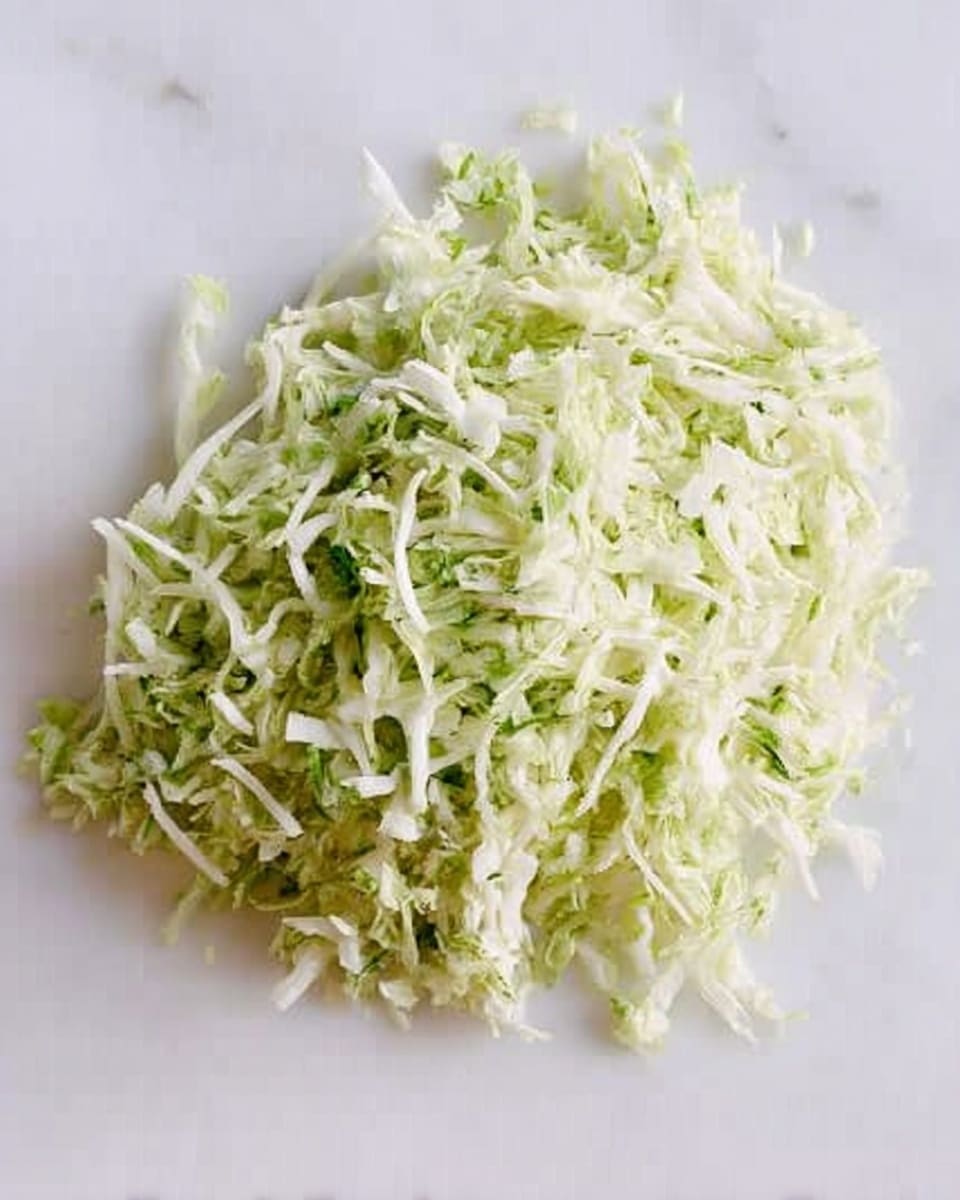 A small pile of shredded light green and white cabbage sits on a white marbled surface. The cabbage strands are thin and uneven, with a fresh, crisp texture visible. There are small bits of darker green cabbage mixed through, creating a subtle contrast. The pile is loosely arranged and looks fresh without any other objects around it. photo taken with an iphone --ar 4:5 --v 7
