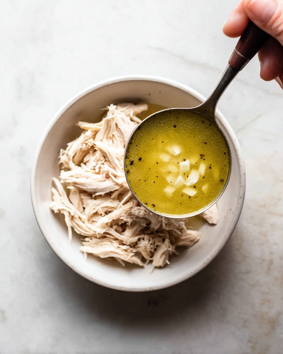 A white bowl is filled with shredded light beige chicken arranged in soft layers. Above the bowl, a ladle holds bright greenish-yellow broth with small white chunks, showing a smooth texture with some visible black pepper specks. A woman's hand holds the ladle by a dark handle over the bowl. The scene is set on a white marbled surface, with soft natural lighting highlighting the textures and colors of the chicken and broth. photo taken with an iphone --ar 4:5 --v 7