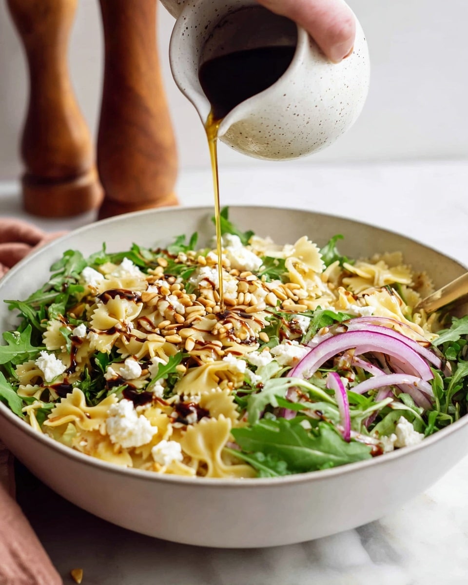 A large white bowl holds a fresh pasta salad with layers of light yellow farfalle pasta, bright green arugula leaves, scattered creamy white crumbled cheese, and thin slices of purple-red onion on the sides. Light brown toasted pine nuts sprinkle the entire dish, and a dark balsamic glaze forms streaks on top. A woman's hand is pouring a thin, shiny golden dressing from a small white speckled ceramic pitcher onto the salad. The bowl sits on a white marbled surface with a wooden pepper mill blurred in the background. Photo taken with an iphone --ar 4:5 --v 7