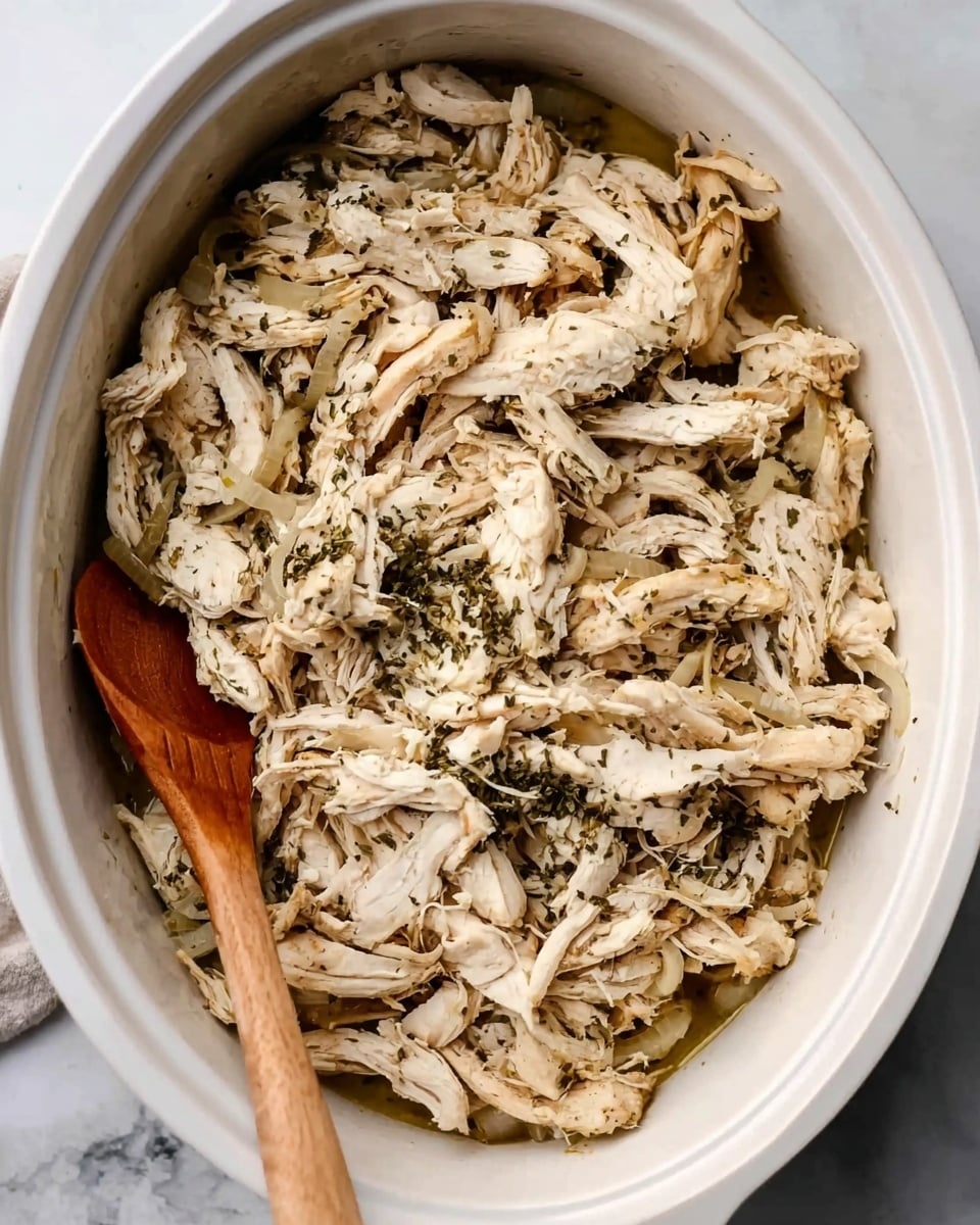 A white oval dish filled with shredded cooked chicken mixed with sliced cooked onions. The chicken is white with some light brown cooked spots and is covered in small green herbs, giving it a speckled look. The dish sits on a surface with a white marbled texture, and a wooden spoon rests inside the dish on the left side. photo taken with an iphone --ar 4:5 --v 7