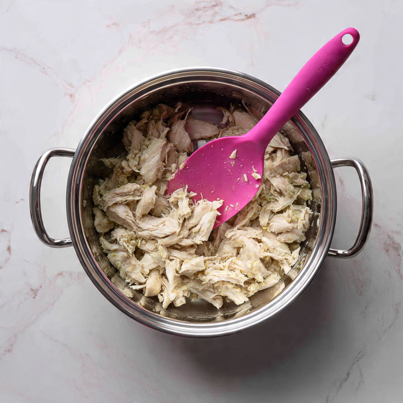 A shiny silver pot filled with many small pieces of pale cooked chicken, slightly steaming, inside the pot is a bright pink spatula resting among the chicken pieces; the pot sits on a white marbled surface with faint gray veins running through it photo taken with an iphone --ar 4:5 --v 7
