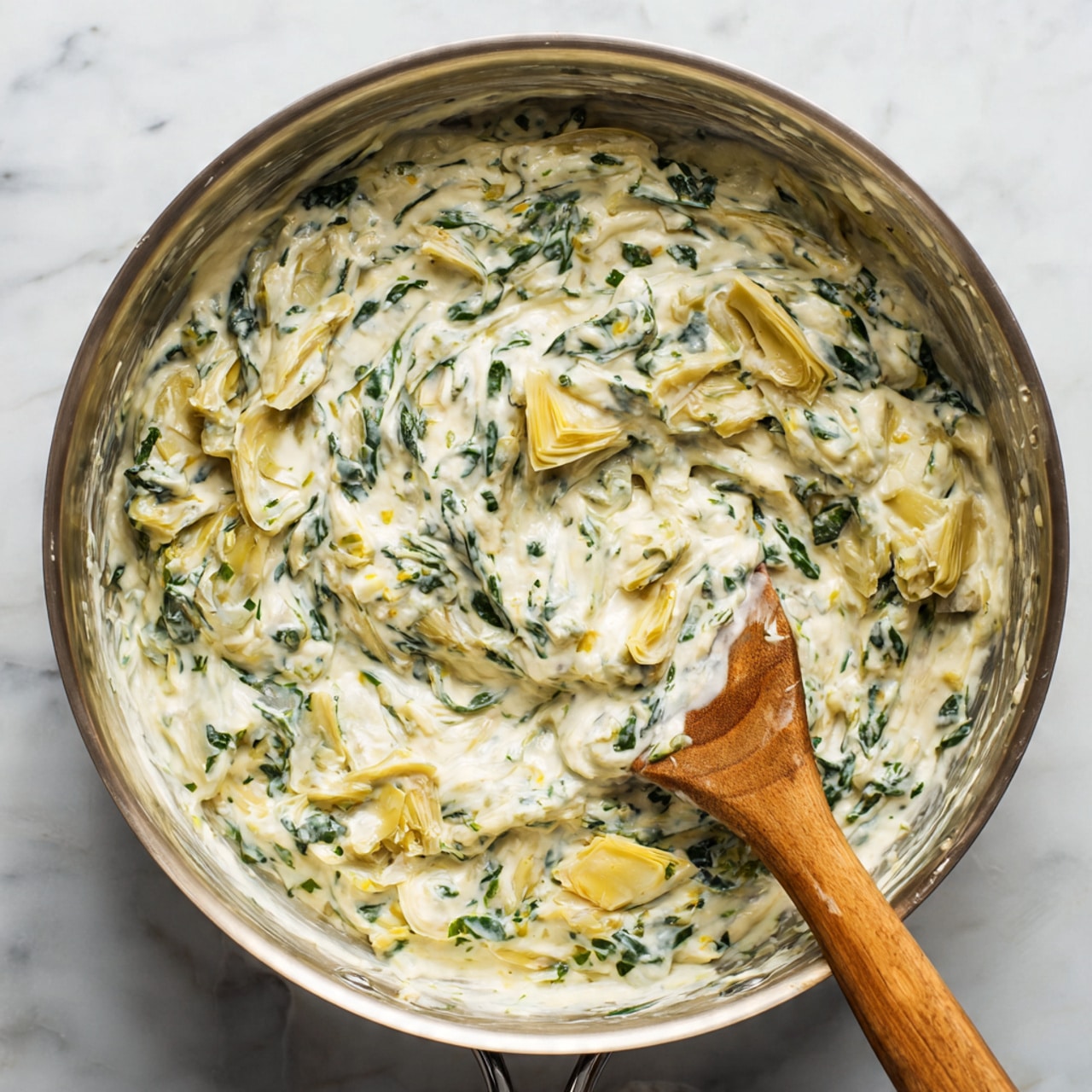 The image shows a thick, creamy spinach and artichoke dip being stirred in a metal pot. The dip has a mostly white base with visible green flecks of chopped spinach mixed throughout, and pieces of light yellow artichoke hearts scattered inside. A wooden spoon is partially covered with the creamy dip, showing the texture to be smooth and thick with some small chunks. The background features a white marbled surface. photo taken with an iphone --ar 4:5 --v 7