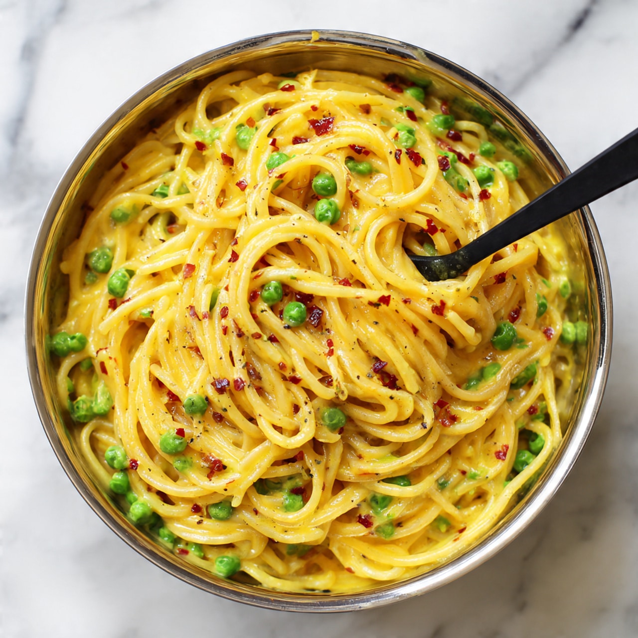A shiny metal bowl filled with creamy yellow pasta mixed with small green peas and tiny red pieces, giving a colorful contrast. The pasta is long and thin, covered in a smooth, thick sauce that looks rich and well-blended. A black spoon is partly submerged in the mixture, with some pasta wrapped around it. The bowl is on a white marbled surface. photo taken with an iphone --ar 4:5 --v 7