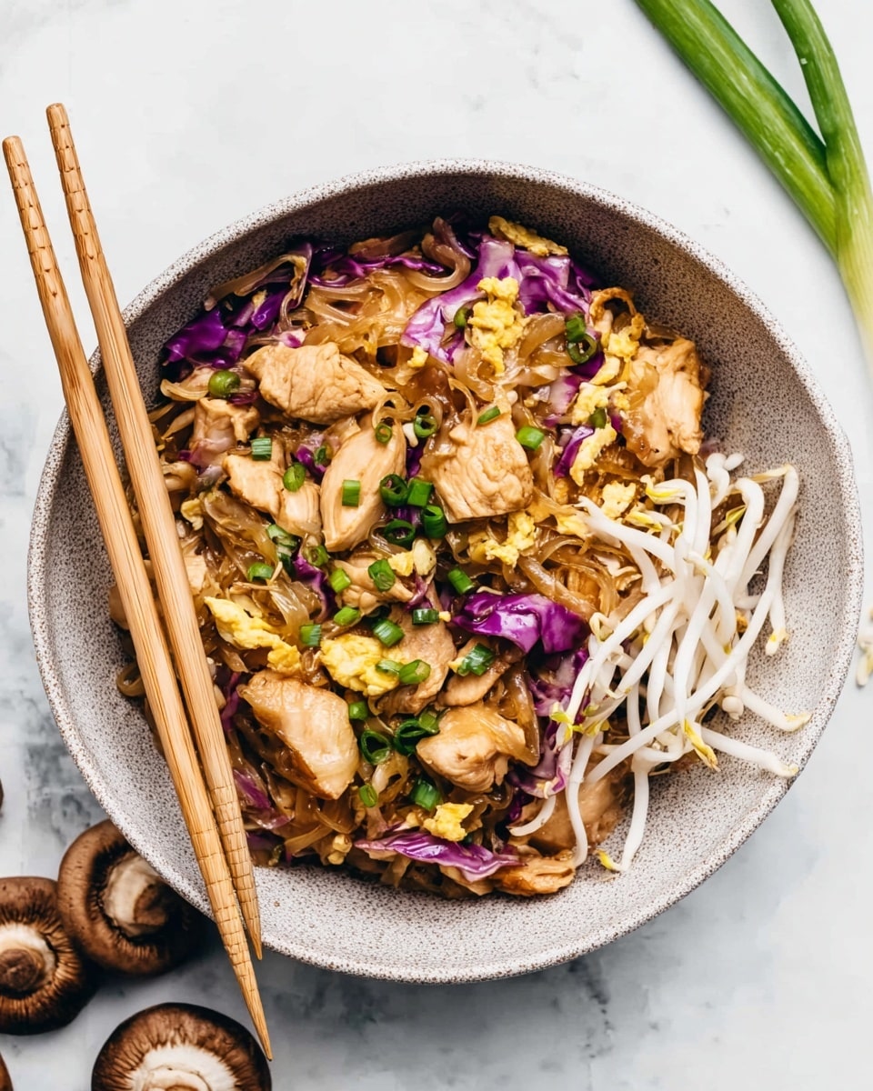 A light grey speckled round bowl holds a colorful mix of stir-fried pieces of light brown chicken layered over cooked translucent brown noodles, with scattered bright purple cabbage pieces, and patches of scrambled yellow egg. Fresh green chopped scallions are sprinkled across the dish, along with some white bean sprouts arranged on the right side. Two light wooden chopsticks rest on the left edge of the bowl, partially inserted into the food. The bowl sits on a white marbled surface with some sliced green scallions and two brown mushrooms nearby. Photo taken with an iphone --ar 4:5 --v 7
