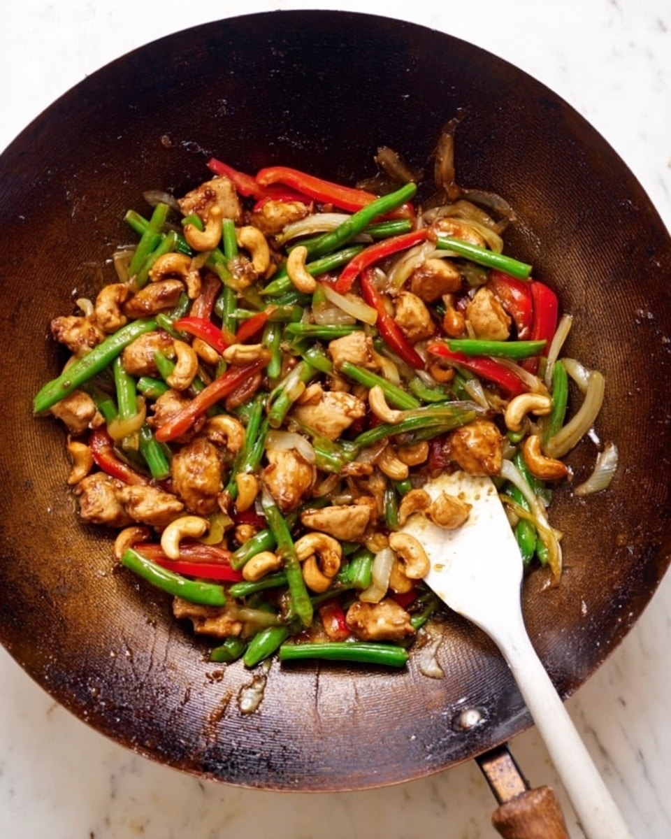A large dark brown wok filled with a colorful stir-fry made of small pieces of cooked chicken, green beans cut in half, thin slices of red bell pepper, and translucent cooked onions. There are also many light brown cashew nuts mixed throughout the dish. A white spatula rests partly inside the wok on the right side, covered slightly with the stir-fry. The wok sits on a white marbled surface, creating contrast with the rich colors of the food. photo taken with an iphone --ar 4:5 --v 7