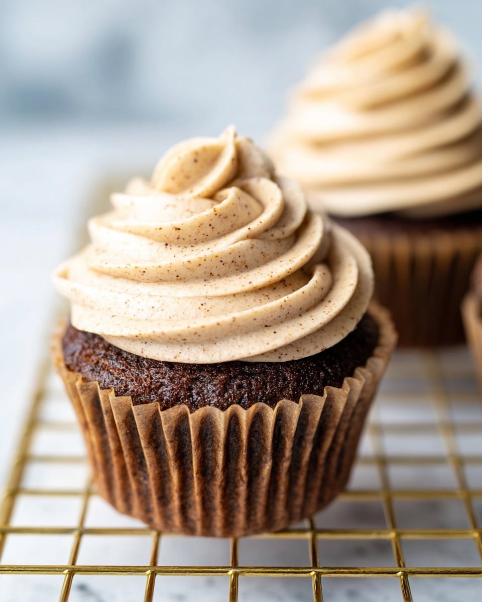 A close-up of a chocolate cupcake with a brown paper liner sitting on a gold wire rack over a white marbled surface. The cupcake has one layer of rich, dark brown cake topped with a thick swirl of light tan frosting that has small brown specks throughout, giving it a textured and creamy look. Behind it, there is a blurred view of another cupcake topped with the same frosting. Photo taken with an iphone --ar 4:5 --v 7