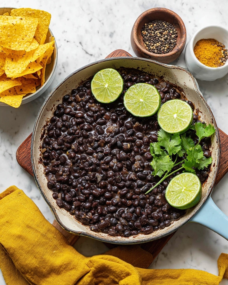 A white bowl filled with dark black beans cooked to a soft texture, topped with small green herb pieces and red chili flakes, with a green lime wedge resting inside the right side of the bowl and a sprig of fresh green cilantro on top; in the background, there are yellow triangular tortilla chips on a white marbled surface and a wooden board, along with a dark wooden bowl holding black peppercorns to the right; the overall scene is bright with a white marbled countertop. photo taken with an iphone --ar 4:5 --v 7