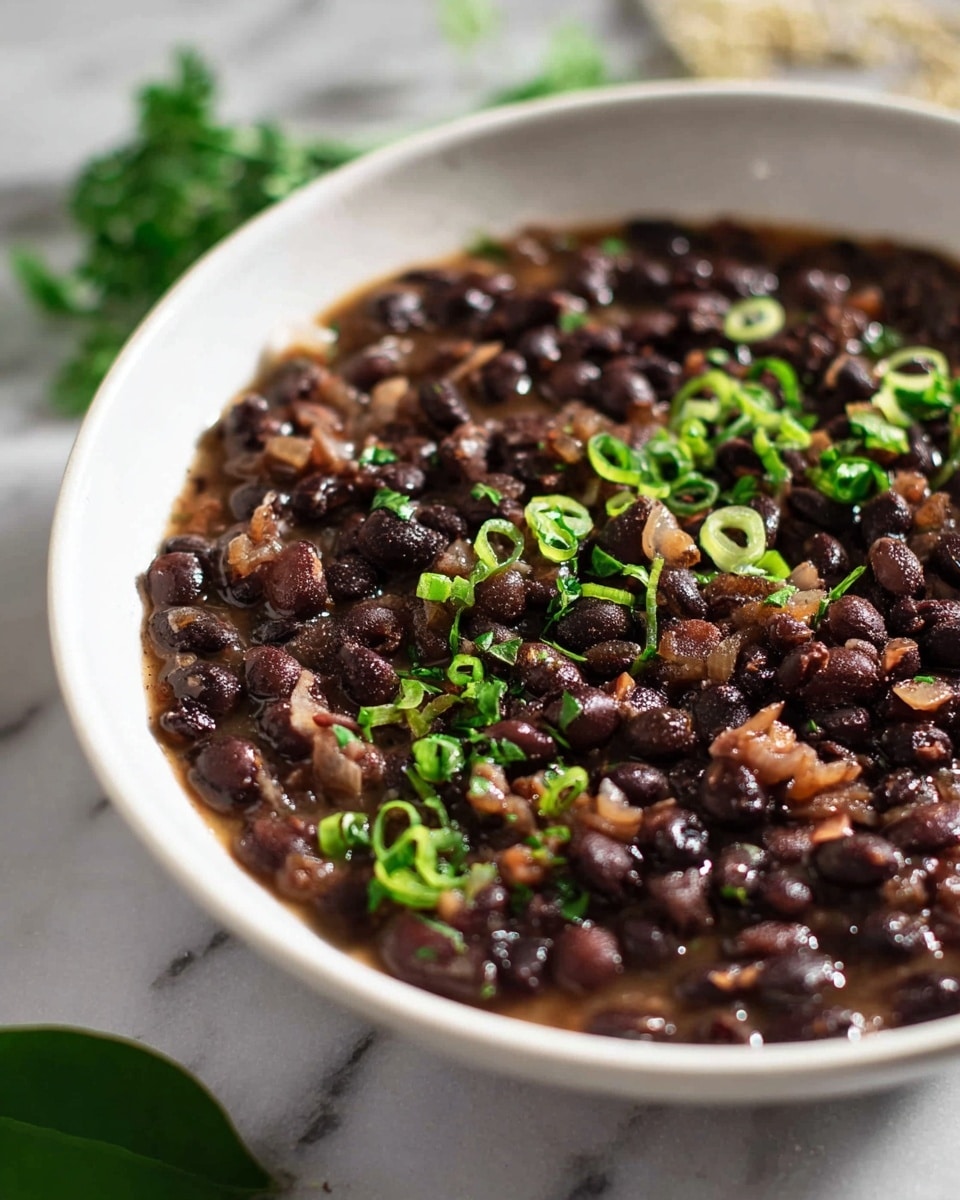 A close-up image of a white bowl filled with black beans cooked with small bits of onions, creating a dark brown and black textured layer with a slightly wet appearance. The beans are topped with thinly sliced green herbs, scattered across the surface, adding a fresh, bright green color contrast. The bowl is placed on a white marbled surface, and there is a single green leaf and some blurred green herbs in the background. The photo taken with an iphone --ar 4:5 --v 7