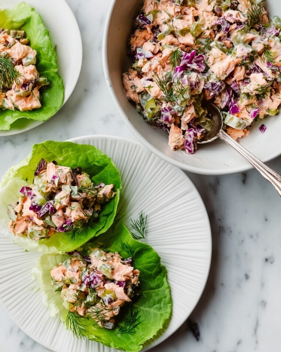 The image shows a white bowl filled with a mix of chopped cooked salmon, green pickles, finely chopped purple cabbage, and fresh dill, all combined in a creamy dressing with a slightly chunky texture. Below, a white plate holds three green lettuce leaves as bases, each topped with a portion of the salmon mixture, displaying a mix of pink, green, and purple colors with creamy and fresh textures. A silver spoon rests inside the bowl on a white marbled surface. Photo taken with an iphone --ar 4:5 --v 7
