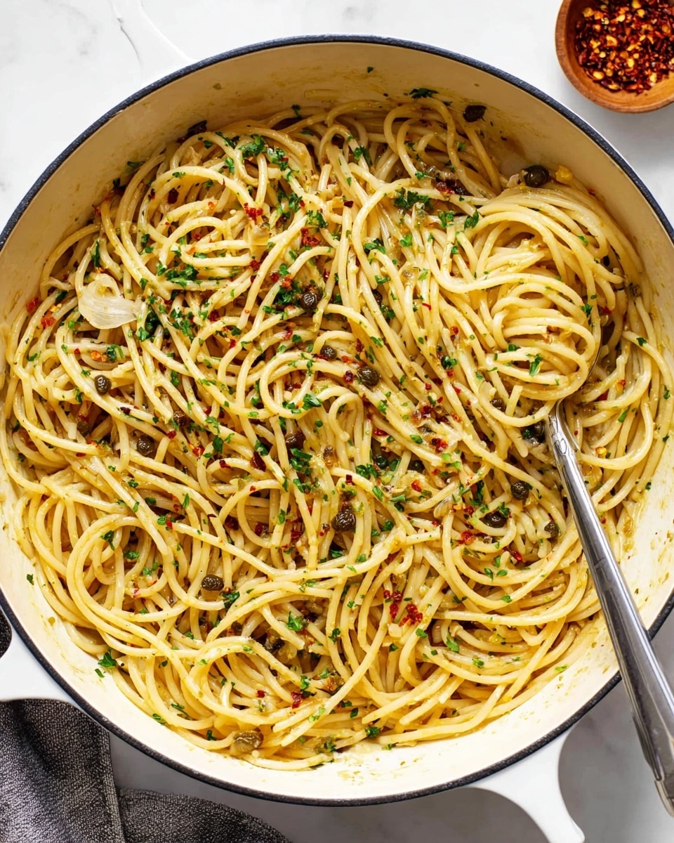 A close-up top view of a white pot filled with spaghetti pasta mixed with thin slices of garlic, small capers, and sprinkled with chopped green herbs and red chili flakes. The spaghetti is light golden yellow, evenly coated with a slight shine of oil. On the right edge, a silver spoon holds a small tangled nest of pasta. The pot sits on a white marbled surface, with a small wooden bowl of red chili flakes partly visible in the top right corner. Photo taken with an iphone --ar 4:5 --v 7