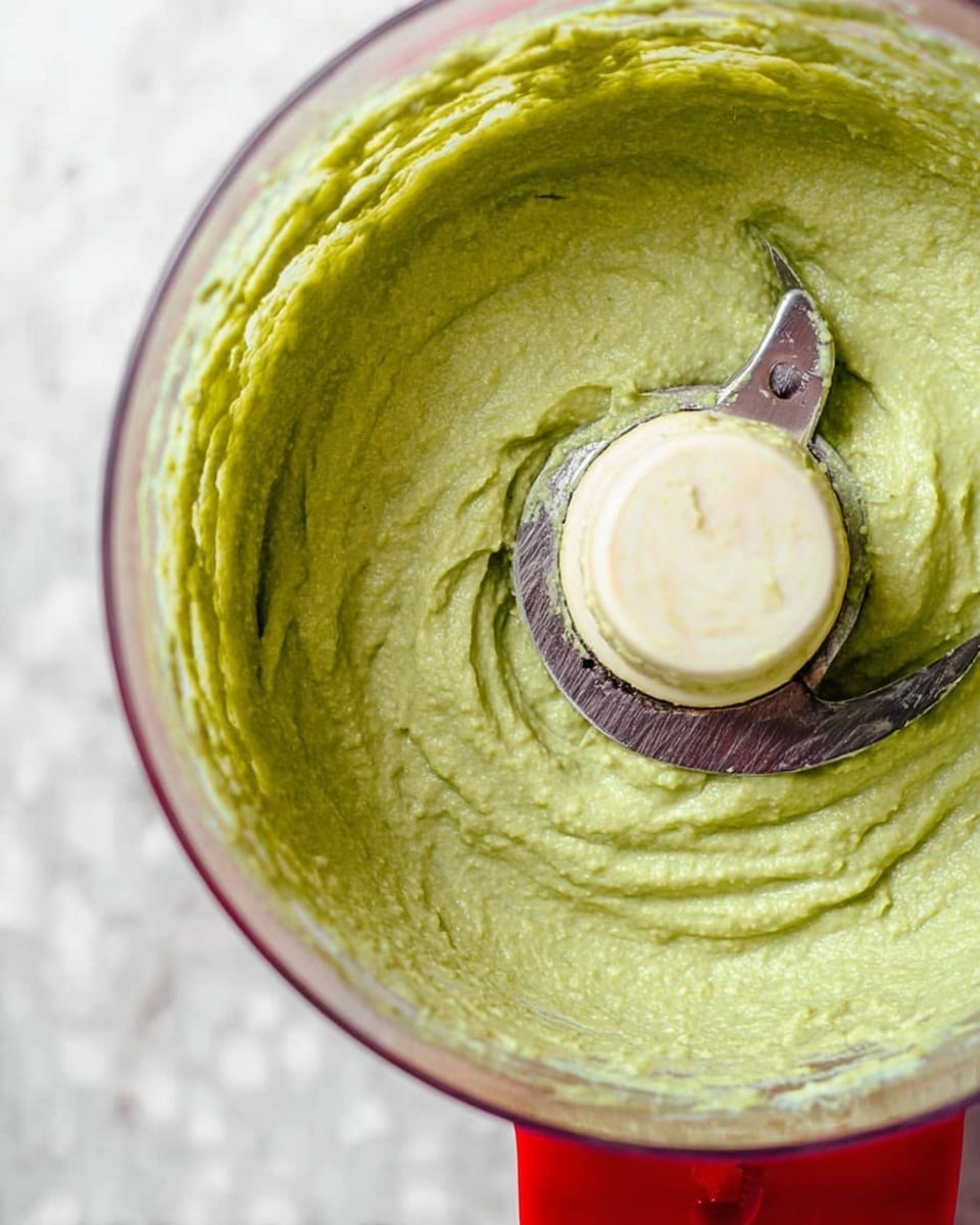 A close-up top view of a food processor bowl filled with smooth, thick green mixture with a creamy texture, spread evenly along the inside walls of the transparent bowl. The metal blade is visible in the center, partly covered by the green mixture. The food processor base is red, and the background surface has a white marbled texture. Photo taken with an iphone --ar 4:5 --v 7