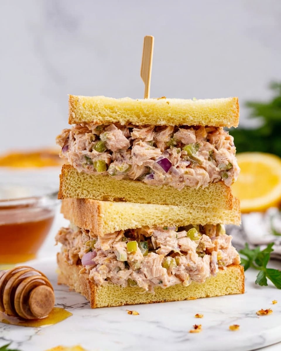 The image shows a sandwich cut into two thick rectangular halves stacked on top of each other. Each half has three layers: the top and bottom layers are soft, light yellow bread with a smooth texture. The middle layer is filled with a chunky mixture of light pink shredded meat, small green pieces of vegetables, and bits of purple onion, giving a colorful and fresh appearance. The sandwich is held together by a wooden skewer stuck through the center. The setting has a white marbled surface with a small bowl of honey and a honey dipper on the left side, some parsley on the right, and blurred background elements. photo taken with an iphone --ar 4:5 --v 7