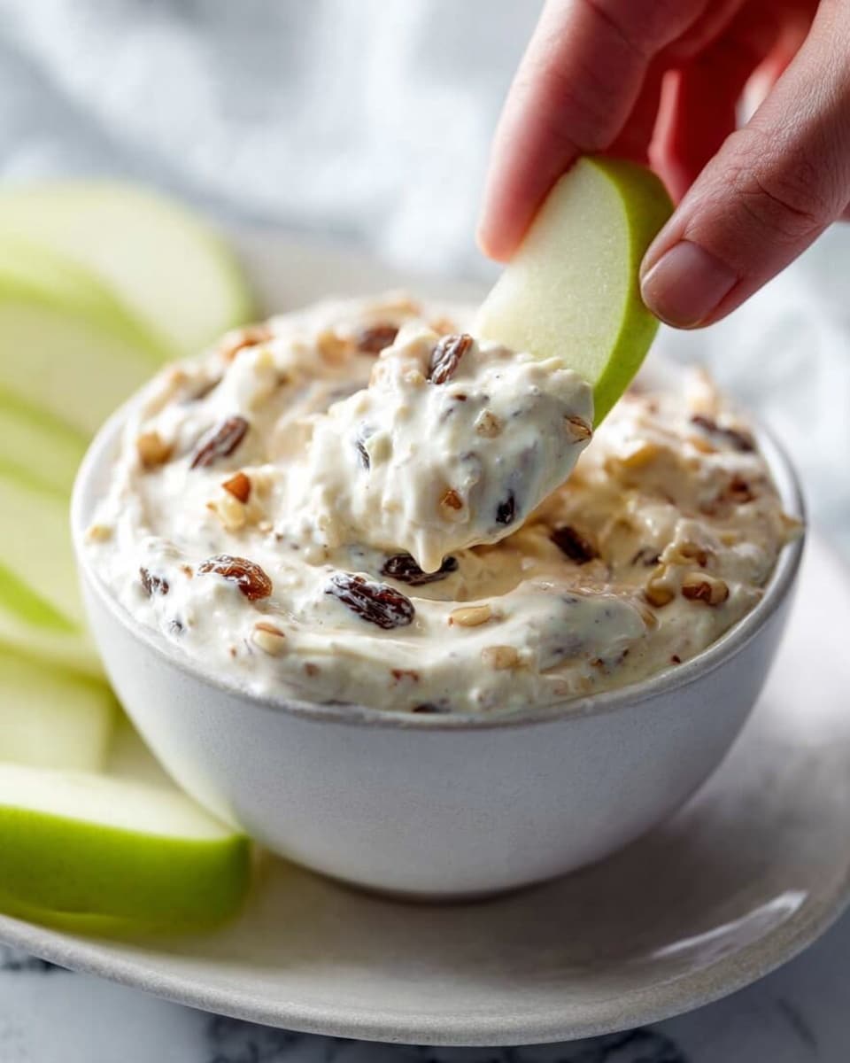 A close-up shot shows a white bowl filled with a creamy dip mixed with dark raisins and small nut pieces, giving it a textured look. A woman's hand is dipping a green pear slice into the thick, white dip, which swirls smoothly around the slice. The bowl sits on a white plate, all placed on a white marbled surface, with additional green pear slices slightly blurred in the background. The lighting is soft and natural, emphasizing the fresh and rich quality of the snack. photo taken with an iphone --ar 4:5 --v 7