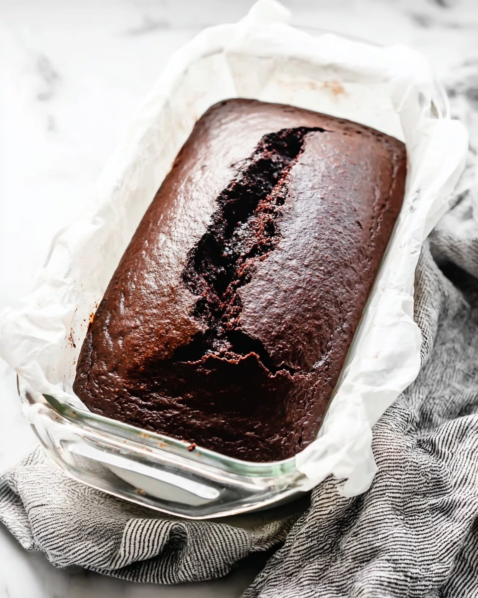 A loaf of dark chocolate cake with a thick, cracked top is resting in a glass Pyrex baking dish lined with white parchment paper. The rich chocolate surface is smooth and shiny, with a deep split running down the middle showing the moist cake inside. The dish sits on a stack of white plates on a white marbled surface, with a gray and white striped cloth nearby. photo taken with an iphone --ar 4:5 --v 7