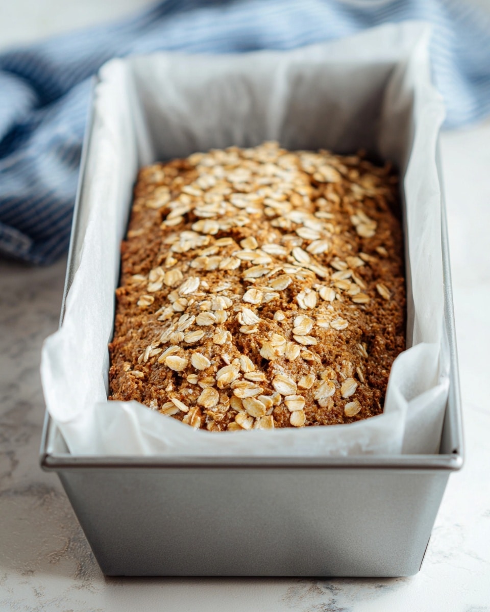 The image shows a brown baked oat loaf inside a silver metal baking pan lined with white parchment paper. The oat loaf has a rough, bumpy texture with many light beige oat flakes spread evenly on the top. The loaf fills the pan almost to the top, and the pan is placed on a white marbled surface. In the background, there is a blurred piece of blue and white cloth. photo taken with an iphone --ar 4:5 --v 7