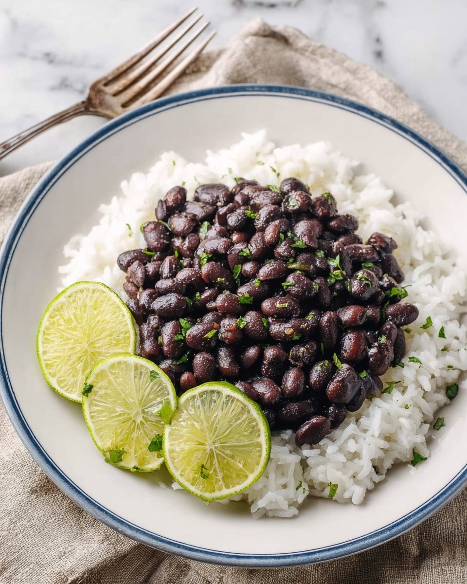 A white plate with a thin blue rim holds a simple dish with two layers. The bottom layer is white rice, fluffy and spread evenly across the plate. On top, there is a mound of shiny black beans sprinkled with small green bits of chopped herbs. To the side, two wedges of bright green lime rest on the rice near the beans. The plate is placed on a beige cloth with lace edges above a white marbled surface, with a silver fork partially visible near the plate. Photo taken with an iphone --ar 4:5 --v 7