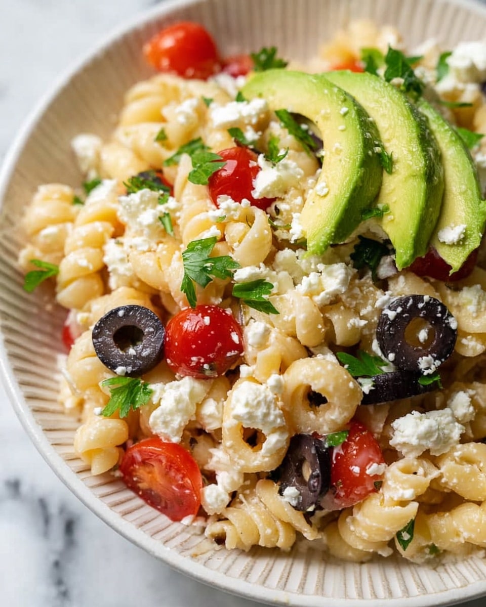 This dish is a close-up view of a pasta salad served in a white bowl with ridged edges. The bottom layer consists of small spiral pasta with a pale yellow color and a slightly creamy texture. Scattered on top are halved cherry tomatoes showing bright red and juicy interiors, black olive slices with smooth, shiny surfaces, and chunks of crumbled white cheese adding a soft, crumbly texture. On one side, there are a few thick, curved slices of green avocado with a creamy appearance, sprinkled with small green bits, likely chopped herbs or peppers. Bright green flat parsley leaves are scattered over the whole dish, adding freshness and contrast. The background shows a white marbled surface. photo taken with an iphone --ar 4:5 --v 7