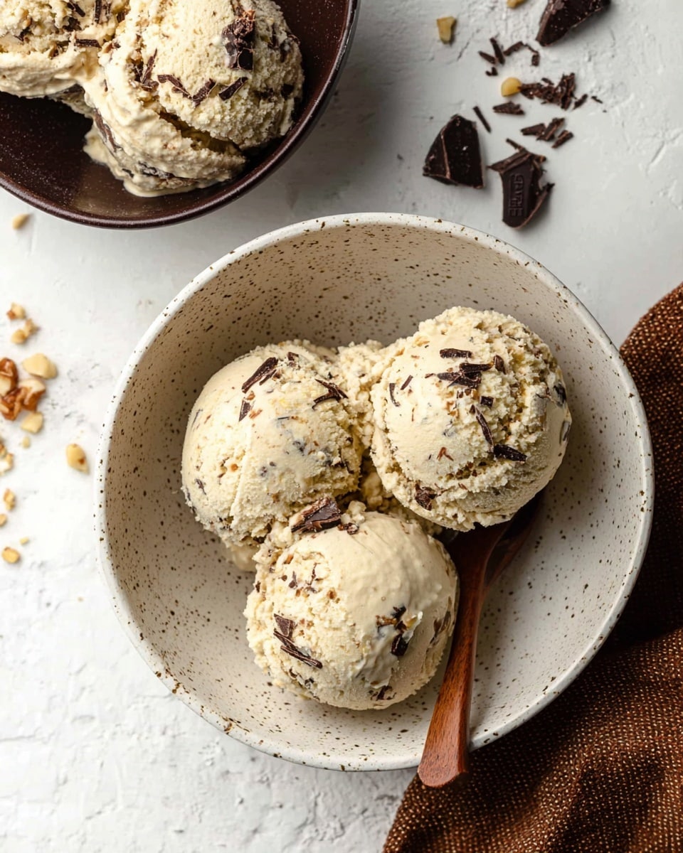 The image shows three scoops of light beige ice cream with visible dark chocolate and small nut pieces mixed inside, placed in a speckled white bowl. The ice cream looks creamy and slightly textured with nut and chocolate bits. A wooden spoon rests inside the bowl beside the scoops. In the top left corner, there is a dark chocolate bowl filled with more scoops of the same ice cream. Small crumbs of chocolate and nuts are scattered around the bowl on a white marbled surface. A brown textured cloth is visible on the right side. photo taken with an iphone --ar 4:5 --v 7