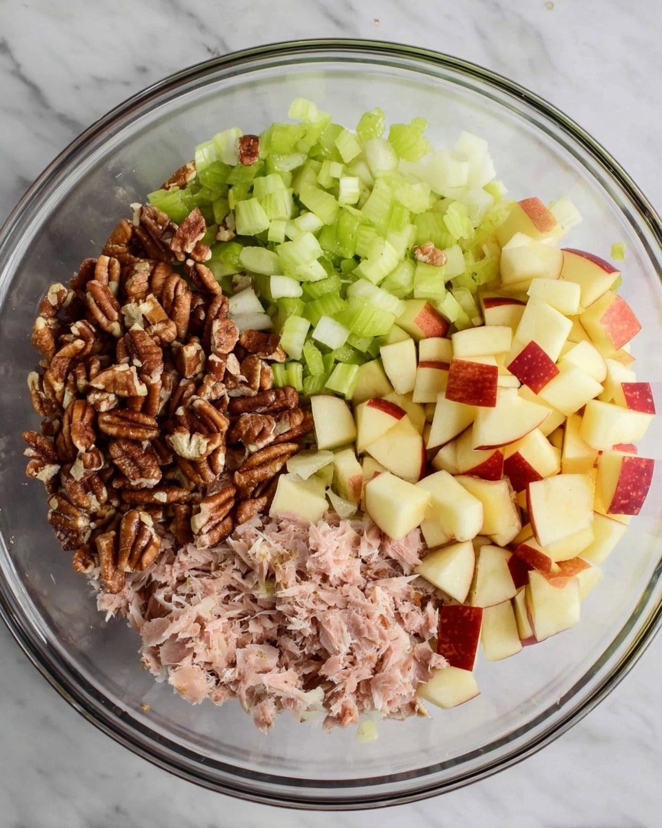 A clear glass bowl holds five different layers of ingredients arranged separately inside it. The first layer is small chopped apple cubes with red skin at the bottom right, showing a light yellow and red mix. Next to it on the left is a layer of chopped pecans, showing rough brown pieces. Above the pecans, there is a pile of chopped green celery. Next to the celery on the right is finely chopped white onion. Lastly, on the bottom right near the onion, there is a layer of light pink shredded tuna. All ingredients show clear texture differences, sitting together in the bowl on a white marbled surface. photo taken with an iphone --ar 4:5 --v 7