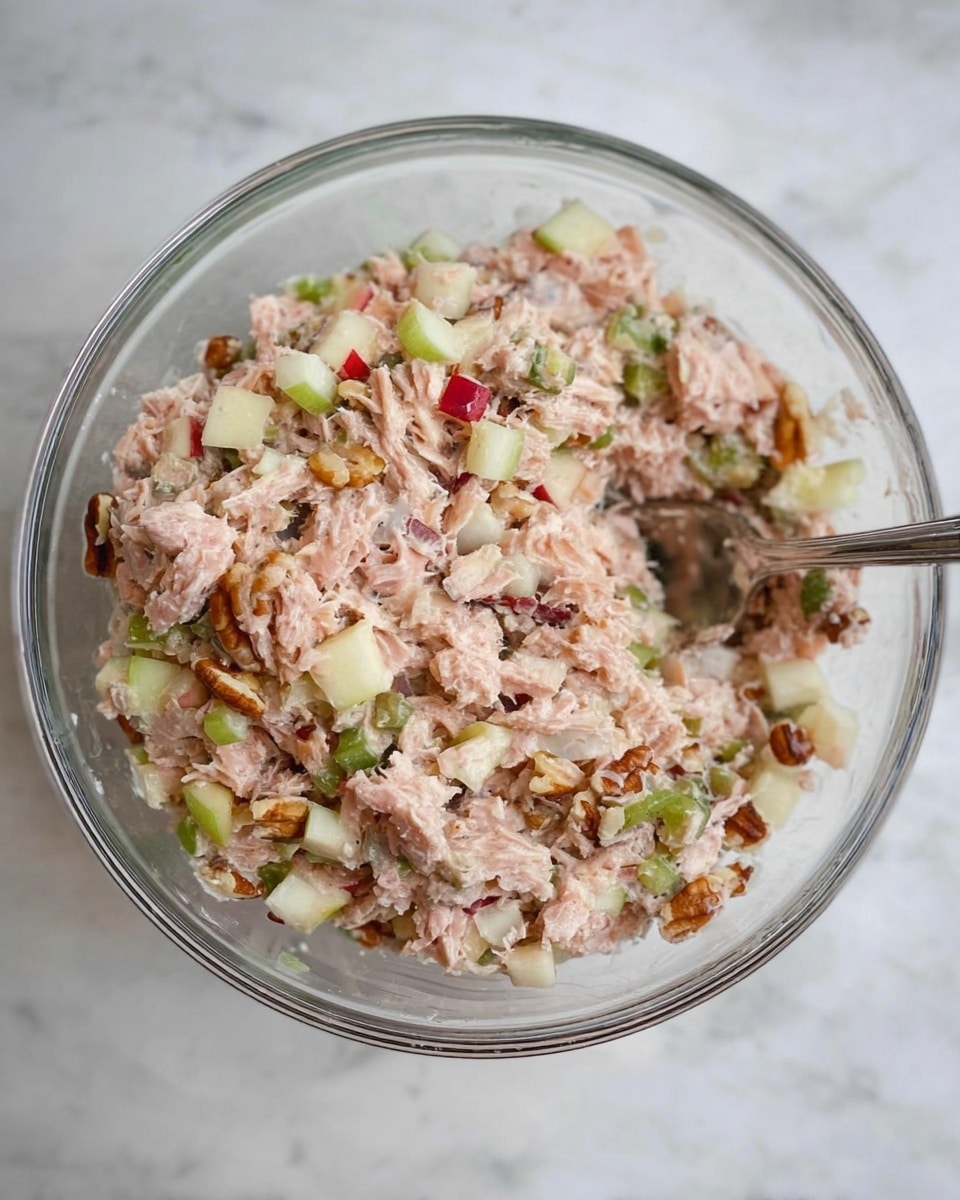 A clear glass bowl filled with a mixture that has about five layers of ingredients visible: shredded light pink tuna, chopped green celery, small cubes of red apple with skin, bits of white onion, and small pieces of brown pecans. The texture looks chunky and moist, with the different colors spread evenly throughout. A silver spoon is partially resting inside the bowl on the right side. The bowl is placed on a white marbled surface. photo taken with an iphone --ar 4:5 --v 7