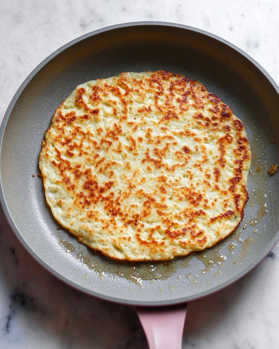 A single round, thick pancake with a golden brown crust and a slightly uneven, bubbly surface sits in the middle of a gray non-stick pan with a pink handle. The pancake's color varies from light beige to golden spots, showing a crispy texture on top and a soft, fluffy inside. The pan rests on a white marbled surface, with some small oil droplets around the pancake, enhancing the cooking effect. photo taken with an iphone --ar 4:5 --v 7