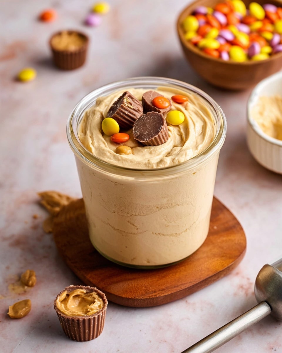 The image shows a clear glass jar filled with a thick, light creamy beige mixture that looks smooth and soft, with visible swirls throughout. On top, there is a small layer of colorful candy pieces including light orange, yellow, and dark brown coated chocolates and halved peanut butter cups with creamy chocolate centers. The jar is placed on a small wooden board, all set on a white marbled surface. Around the jar, there are a few small peanut butter cups scattered and a wooden bowl filled with more colorful coated candies in the background, along with a white bowl holding more of the creamy beige mixture. A metal scooping tool lies to the right of the jar, partially visible. The photo taken with an iphone --ar 4:5 --v 7