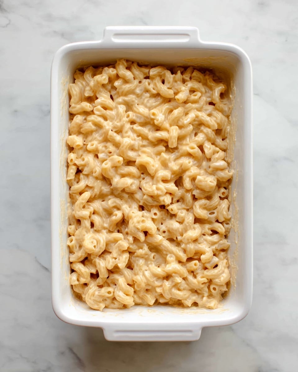 A white rectangular baking dish filled with one layer of macaroni pasta coated in a creamy, pale yellow cheese sauce, evenly spread so the noodles are fully covered. The dish is placed on a white marbled surface with a clean, simple look. photo taken with an iphone --ar 4:5 --v 7