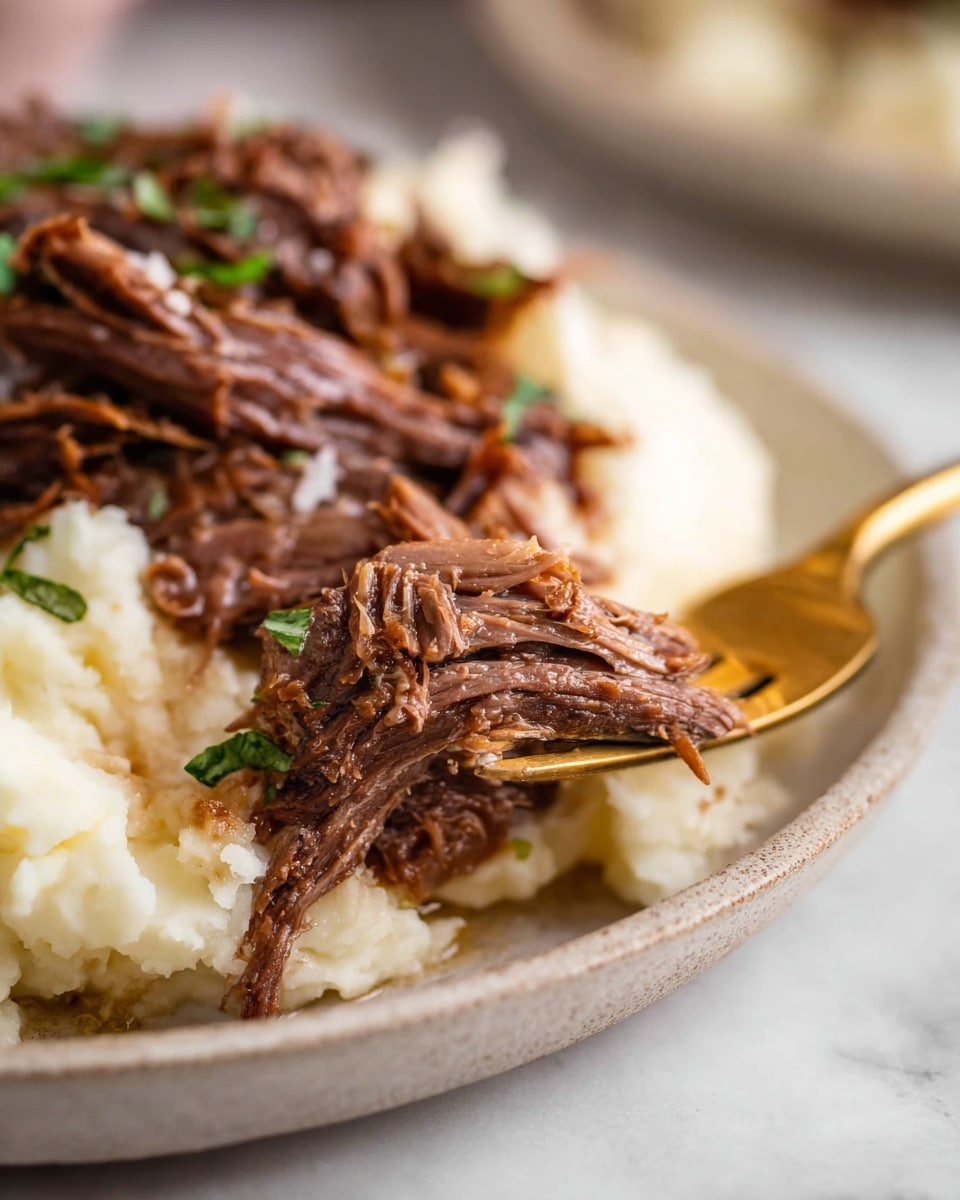 The image shows a close-up of tender, shredded brown meat placed on top of creamy off-white mashed potatoes. The meat has a juicy texture with visible strands and some green herb garnishes scattered on it. Part of a shiny gold fork is underneath the meat, holding a bite-size piece. The food is served on a white plate with a soft focus on the rest of the mashed potatoes in the background. The overall scene is set on a white marbled surface. Photo taken with an iphone --ar 4:5 --v 7