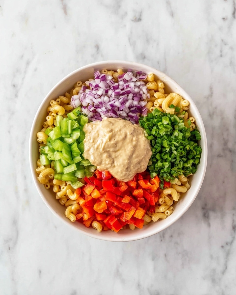 A white bowl sits on a white marbled surface filled with four distinct layers: the bottom layer is light yellow elbow macaroni filling most of the bowl; on top of the pasta, there are four sections of finely chopped vegetables arranged in a circle – bright green celery on the left and right sides, red bell pepper at the bottom right, and purple onion at the top center; in the middle of the bowl, on top of the vegetables and pasta, there is a dollop of thick, creamy beige dressing. Photo taken with an iphone --ar 4:5 --v 7