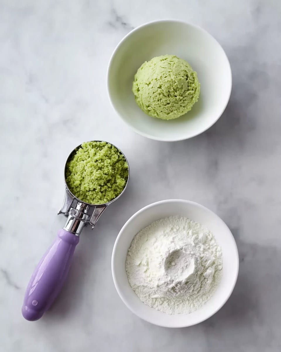 A white bowl in the middle top contains one green round scoop of a textured mixture. Below it, another white bowl holds a white powder with an indentation in the center, showing the shape of a pressed round item. To the left of the bowls, a purple ice cream scoop with metal parts holds a green round scoop of the same textured mixture. All items rest on a white marbled surface. photo taken with an iphone --ar 4:5 --v 7