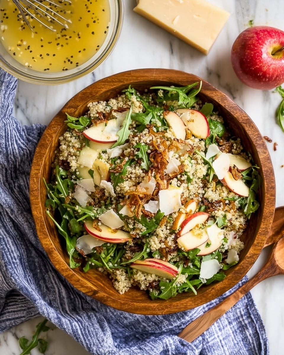 A wooden bowl filled with a colorful quinoa salad layered with white quinoa grains as the base, bright green arugula leaves scattered throughout, thin slices of red and white apple pieces, and topped with light brown crispy fried shallots and white cheese shavings. The bowl sits on a white marbled surface with a striped blue and white cloth partially visible underneath. Nearby, there is a wedge of cheese, a whole red apple, and a glass bowl filled with a light yellow dressing with black pepper specks and a metal whisk inside. A wooden spoon is placed beside the bowl. Photo taken with an iphone --ar 4:5 --v 7
