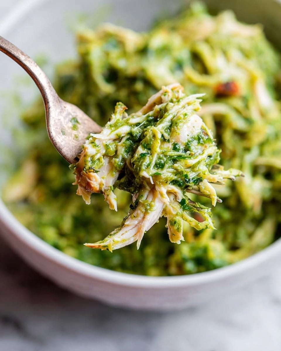The image shows a close-up of a fork holding a serving of shredded chicken mixed with a bright green sauce that looks creamy and textured, with many small bits of herbs or vegetables visible. The chicken is mostly white with some browned edges. The background includes a white bowl filled with the same green chicken mixture sitting on a white marbled surface. The colors are fresh with green and light brown tones. photo taken with an iphone --ar 4:5 --v 7