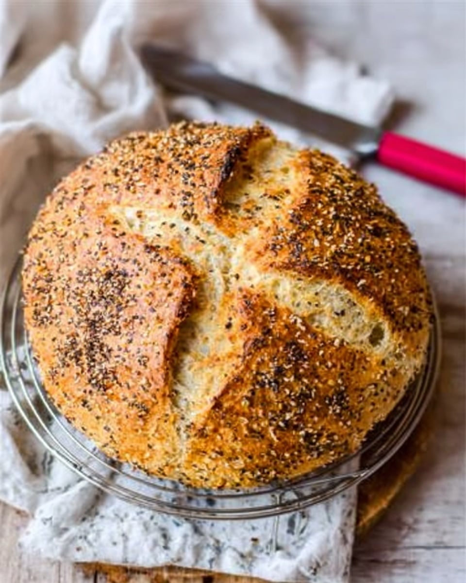 A round loaf of bread with a golden brown crust is placed on a silver wire cooling rack. The top of the bread is covered with mixed seeds and grains, giving it a rough texture. The loaf has a cross-shaped cut on top showing a light, soft inside. To the side, a knife with a red handle rests partly on the edge of a white marbled surface, and a white cloth with a subtle pattern is slightly visible in the background. photo taken with an iphone --ar 4:5 --v 7