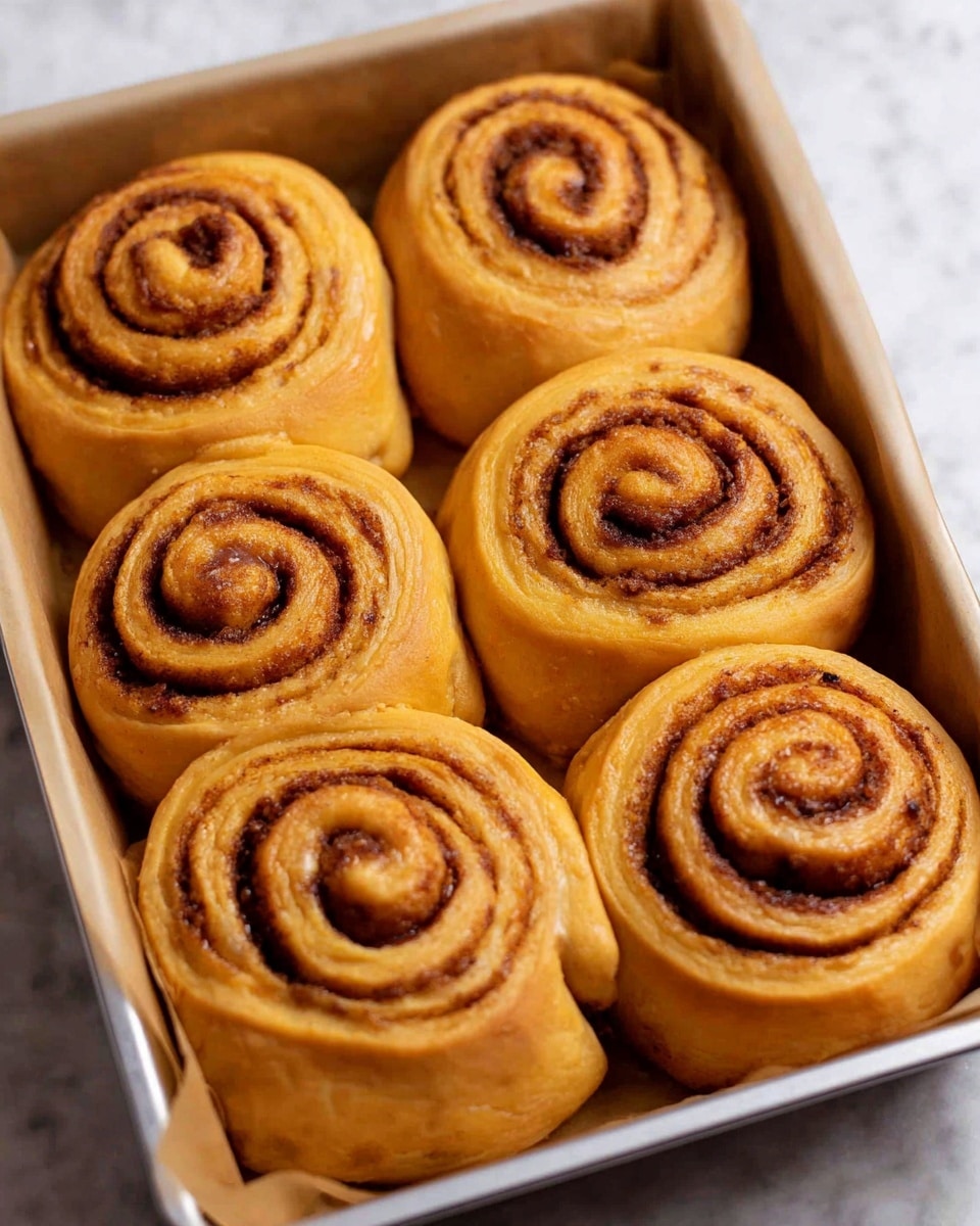 The image shows nine golden-brown cinnamon rolls in a silver baking tray, arranged in three rows of three. Each cinnamon roll has a tight spiral shape with visible layers of dough and cinnamon filling that create a dark brown swirl pattern inside the lighter golden dough. The outer edges of the rolls are smooth and slightly glossy, and they sit closely together but not touching. The tray is lined with light beige baking paper, and the background is a white marbled surface. photo taken with an iphone --ar 4:5 --v 7